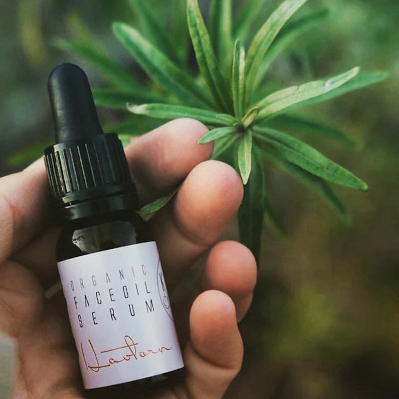 KALIFLOWER ORGANICS Gesichtsöl für empfindliche/trockene Haut, Sanddorn. Hand mit Flasche vor einer Pflanze. 